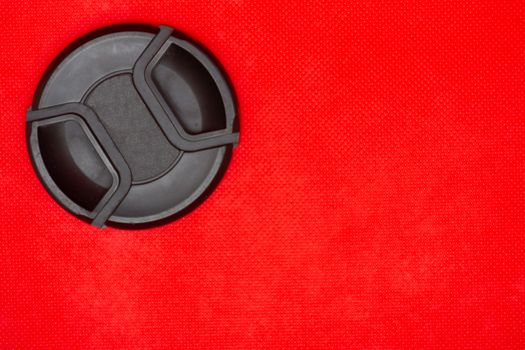 Close up of a black circle lens cap top left corner for DSLR camera lens on a rich red background.