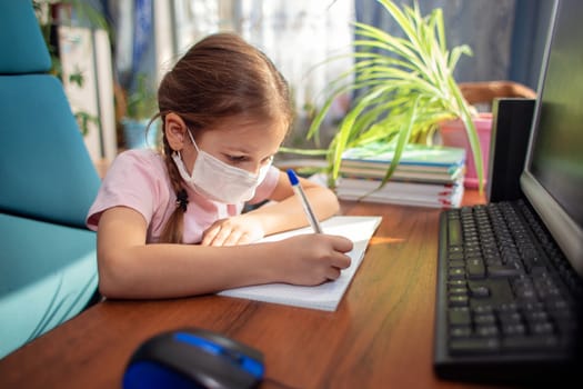 Girl schoolgirl in a medical mask sits at home in quarantine and does homework in a notebook