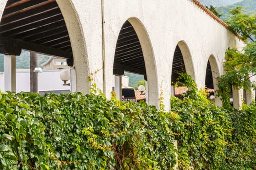 A white stone building with arches and green bushes and ivy planted near it.