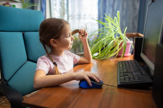 Girl schoolgirl with pigtails sits at home in an armchair at a desktop computer and drives a computer mouse