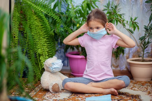 A girl puts a medical mask on herself, next to her her teddy bear is also wearing a mask during quarantine