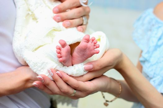 Mom and dad hands hold small legs of their newborn baby, wrapped in a white warm blanket.
