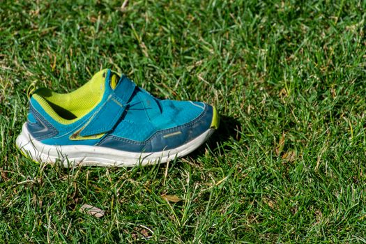 Child's blue and green shoe sits in the sunshine on the green grass prepared for activity and sports in the summer.