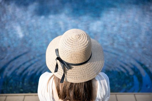 Back view of relaxing woman in swimsuit and straw hat  sitting near the swimmingpool