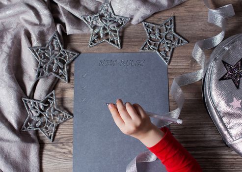 The child writes new rules for the new year on gray paper. New plan, words, message. Motivation, schedule, task, new life. Christmas, holiday. The silver background. Snowflakes and Christmas toy