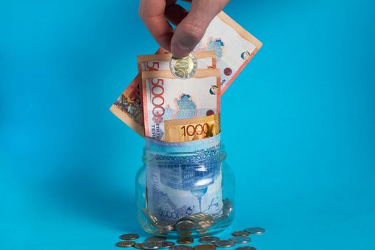 The hand puts a tenge coin in a glass jar with money. Kazakhstan. KZ. KZT. Banknote, paper currency, coins, Bank. Salary, credit, savings, pay and spend. Business and Economics.