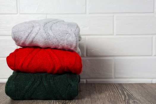 Clothing, fashion, style. A stack of multicolored sweaters are placed on a wooden table near a white brick wall. Hygge, cozy, cashmere, soft. Home routine, order, storage of things.