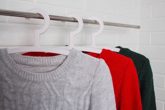Clothes hang on hangers. Sweaters in red, green and melange colors. Fashion and style, showcase, store, concept. White brick wall, comfort and cleaning, design, hygge, wardrobe. and knitwear. Close up