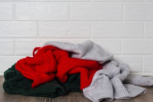 A pile of clothes against a white brick wall. Fashion, style. Sweaters in red, green and melange colors. Mess, home routine, storage. Wardrobe, hygge and knitwear.