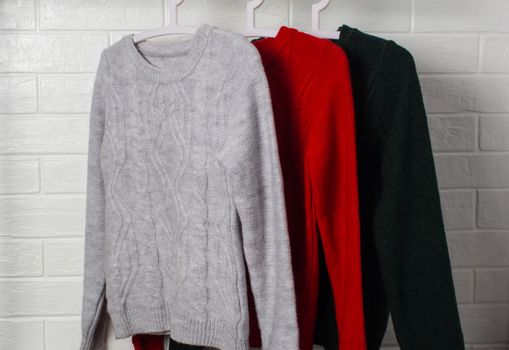 Clothes hang on hangers. Sweaters in red, green and melange colors. Fashion and style, showcase, store, concept. White brick wall, comfort and cleaning, design, hygge, soft and knitwear. Wardrobe.