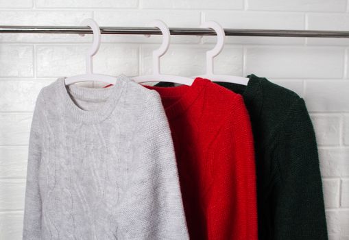 Clothes hang on hangers. Sweaters in red, green and melange colors. Fashion and style, showcase, store, concept. White brick wall, comfort and cleaning, design, hygge, soft and knitwear. Wardrobe.