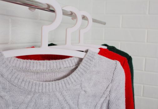 Clothes hang on hangers. Sweaters in red, green and melange colors. Fashion and style, showcase, store, concept. White brick wall, comfort and cleaning, design, hygge, wardrobe. and knitwear. Close up