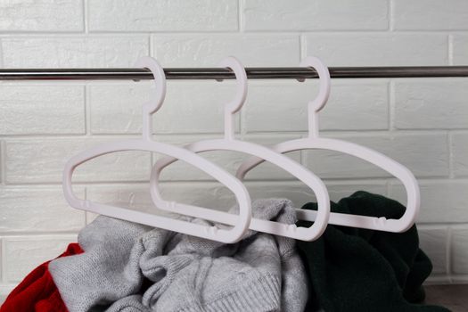 Empty white hangers, and under them a pile of clothes. Fashion, style. Sweaters in red, green and melange colors. White brick wall. Mess, home routine, storage. Wardrobe, hygge and knitwear.