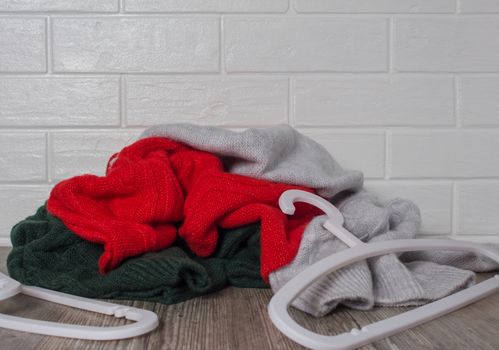 A pile of clothes against a white brick wall. Scattered clothes hangers. Fashion, style. Sweaters in red, green and melange colors. Mess, home routine, storage. Wardrobe, hygge and knitwear.	