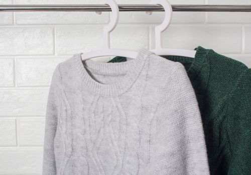 Clothes hang on hangers. Sweaters green and melange colors. Fashion and style, showcase, store, concept. White brick wall, comfort and cleaning, design, hygge, soft and knitwear. Wardrobe.