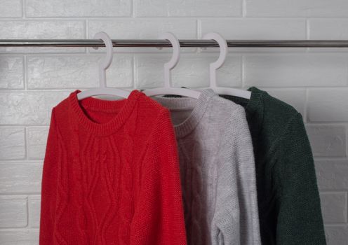 Clothes hang on hangers. Sweaters in red, green and melange colors. Fashion and style, showcase, store, concept. White brick wall, comfort and cleaning, design, hygge, soft and knitwear. Wardrobe.
