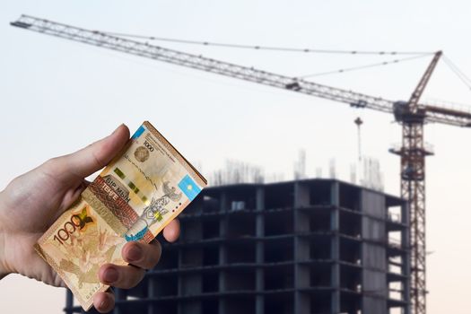 Hands holding money tenge on the background of the house under construction. Buying a new apartment in Kazakhstan for tenge. New Buildings In Kazakhstan. Real estate services and Bank loans.