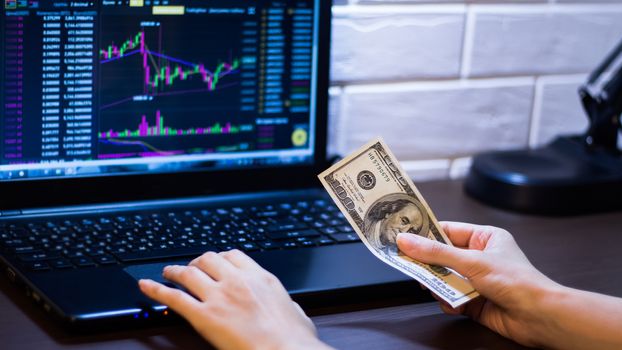 A trader trades stocks,bonds and securities or currencies on the Forex market or stock exchange for us dollars.Hands holding a dollars in front of a laptop monitor with a price chart.Forex and trading