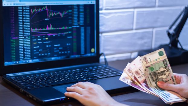 Hands holding Russian rubles in front of a computer monitor with the currency market and Forex trading chart. Trading stocks, bonds, and securities on the stock exchange. Trader in Russia with ruble.