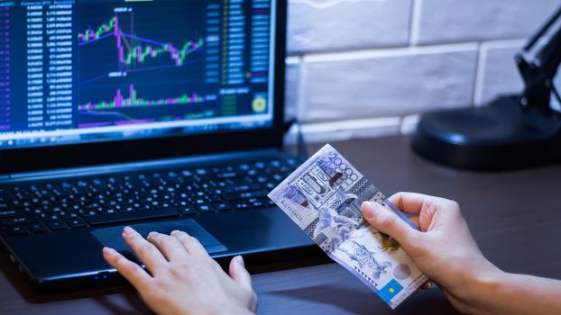 Tenge in Kazakhstan against the background of a laptop with an open chart of the currency market or stock exchange. Hands holding Tenge in front of the computer. Tenge KZT, bank of Kazakhstan.