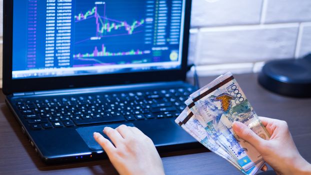 Tenge in Kazakhstan against the background of a laptop with an open chart of the currency market or stock exchange. Hands holding Tenge in front of the computer. Tenge KZT, bank of Kazakhstan.