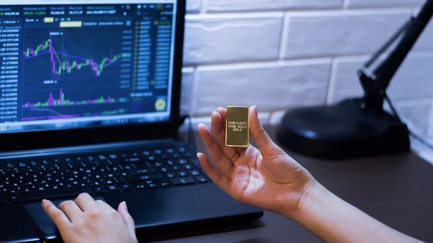 The hand holds a gold bar on the background of a laptop on which there is a trading price chart. One ounce of gold in hand. Trading gold on the stock exchange and Forex.