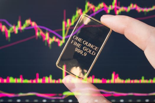 The hand holds a gold bar on the background of a laptop on which there is a trading price chart. One ounce of gold in hand. Trading gold on the stock exchange and Forex.