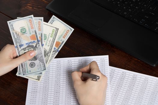 Work in the office with finance. A cashier holds money dollars over an office work space with documents, a cash register, a phone, and a computer. Obligation to pay wages and debts in the company.