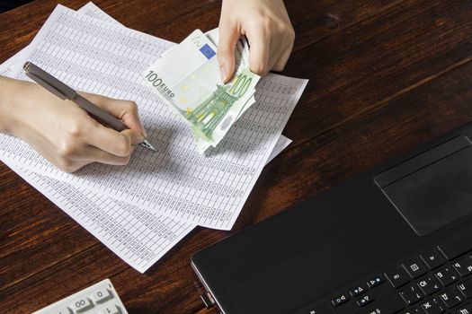 Obligation to pay wages and debts in the company.A cashier holds money Euro over an office work space with documents, a cash register,a phone,and a computer.Work in the office with finances in Europe