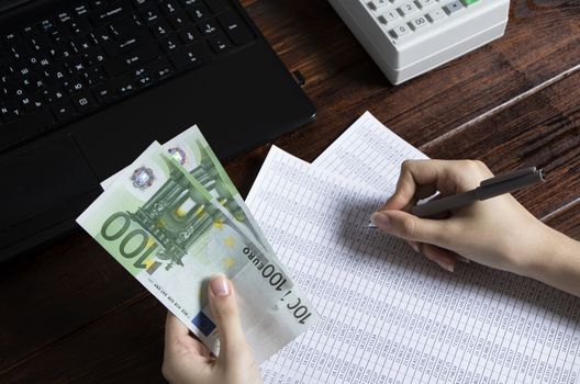 Obligation to pay wages and debts in the company.A cashier holds money Euro over an office work space with documents, a cash register,a phone,and a computer.Work in the office with finances in Europe