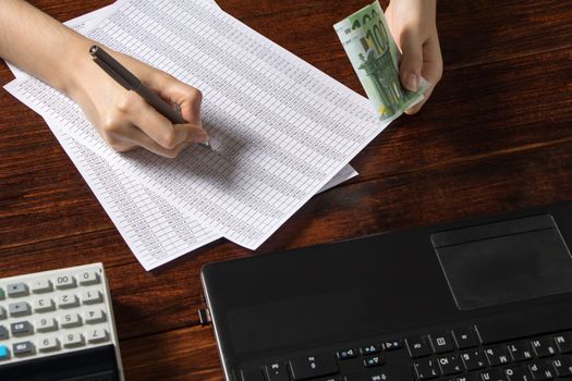 Obligation to pay wages and debts in the company.A cashier holds money Euro over an office work space with documents, a cash register,a phone,and a computer.Work in the office with finances in Europe