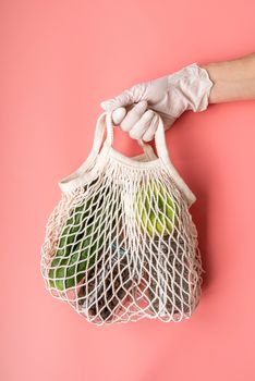 Coronavirus food supplies concept. Food delivery. Hand in a glove holding white eco friendly mesh bag with food supplies on pink background