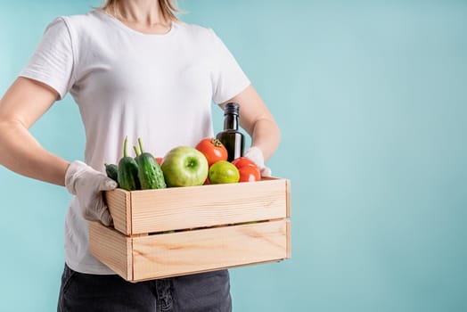 Coronavirus food supply. Woman in gloves holding a wooden box full of vegetables isolated on blue background with copy space