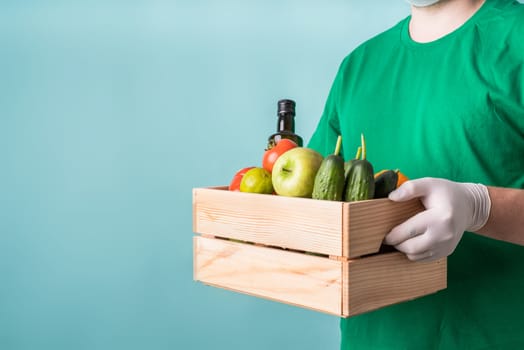Coronavirus food supply. Man in gloves holding a wooden box full of vegetables isolated on blue background with copy space