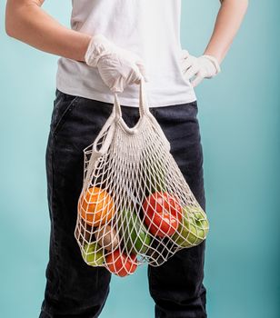 Zero waste concept. Coronavirus food delivery. Woman in gloves holding a white mesh bag with vegetables isolated on blue background