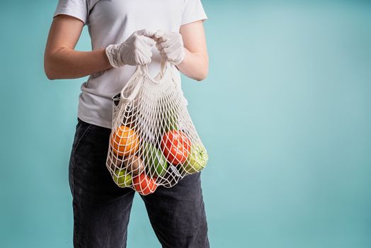 Zero waste concept. Coronavirus food delivery. Woman in gloves holding a white mesh bag with vegetables isolated on blue background with copy space