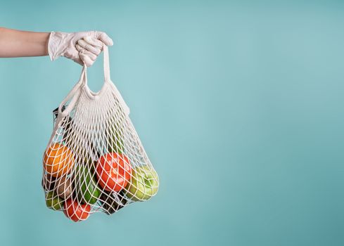 Zero waste concept. Coronavirus food delivery. Woman in glove holding a white mesh bag with vegetables isolated on blue background with copy space