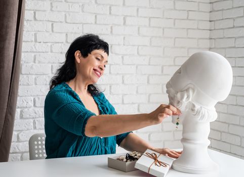 Beautiful smiling middle aged woman trying on the jewelery on the mannequin sitting at the table