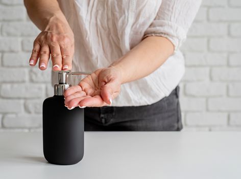 Middle aged woman hands using sanitizer gel pump dispenser on white brick wall background. Copy space