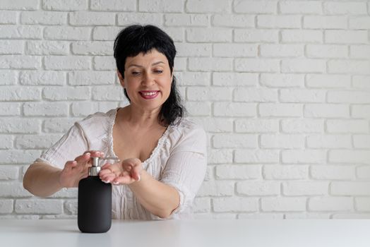 Middle aged woman hands using sanitizer gel pump dispenser on white brick wall background. Copy space