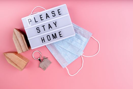 Stay home. Self quarantine. The words Please Stay Home on a lightbox with wooden house and masks top view on pink background