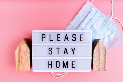 Stay home. Self quarantine. The words Please Stay Home on a lightbox with wooden house and masks top view on pink background