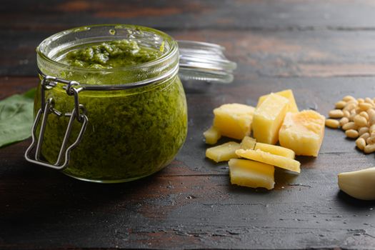 Green Pesto Basil Sauce in glass jar closeup. with ingredients parmesan basil pine nuts on dark wood table planks vintage side view.