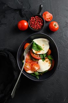 Cherry tomatoes, mozzarella buffalo cheese, basil, pesto sauce, and spices on black slate stone chalkboard. Italian traditional caprese salad ingredients. Mediterranean food in black bowl with fork.