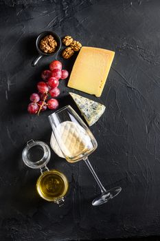 Cheese appetizer selection or whine snack set. Variety of mold french cheese, grapes, pecan nuts, white sauvignon wine and honey over black backdrop, top view, copy space vertical.