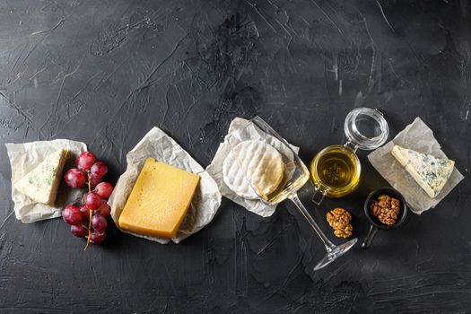 Cheese appetizer selection or whine snack set. Variety of mold french cheese, grapes, pecan nuts, white sauvignon wine and honey over black backdrop, top view, copy space.