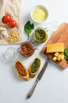 Glass jars with red and green pesto and cooking ingredients Parmesan cheese, basil leaves, pine nuts, white stone background, close up top view. vertical