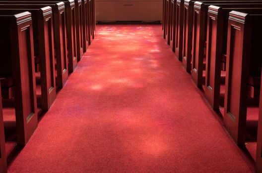 Red carpeted church aisle between pews illuminated by light from stained glass window