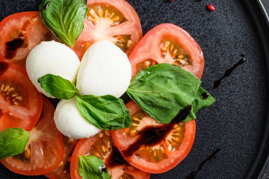 Caprese salad Tomato and mozzarella slices with basil leaves on olive wood dark table top view close up on black plate.