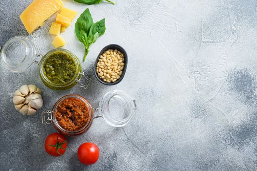 Glass jars with red and green pesto and cooking italian recipe ingredients Parmesan cheese, basil leaves, pine nuts, olive oil, garlic, salt, tomatoes top view on grey concrete surface space for text.